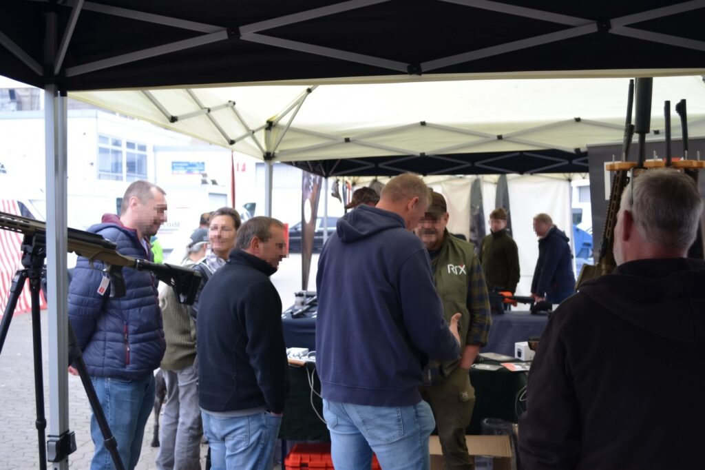 Besucher unter einem Ausstellerzelt, links im Bild ein Gewehr auf einem Stativ.